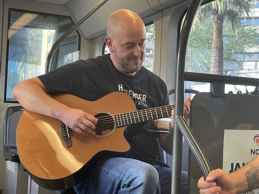 Bald man in black shirt plays acoustic guitar while sitting on the light rail