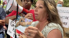 Kristina Salinas speaks into a bullhorn at a protest, with people holding signs behind her. She's holding up a photo of her sister Kelly Masten's bruised legs, propped up in a hospital bed.