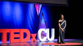 Large letters on stage say TEDxCU. A person stands to the right in a large black gown.