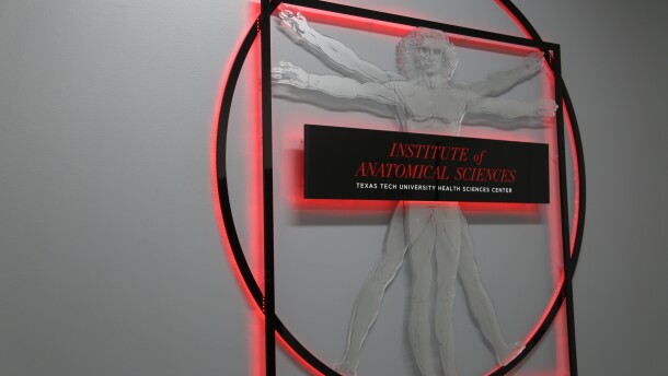 A sign showing Leonardo da Vinci's The Vitruvian Man, with text that reads 'Institute of Anatomical Sciences - Texas Tech University Health Sciences Center.' It has red lights glowing from behind it and onto the white wall.