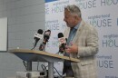 Sorensen wears a tan suit and blue shirt and stands at a podium. 