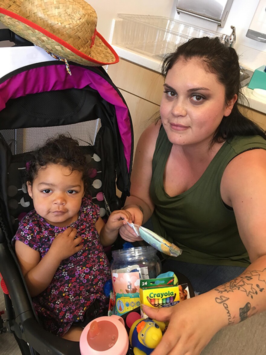 A toddler in a pink shirt sits in a stroller with her mother by her side.