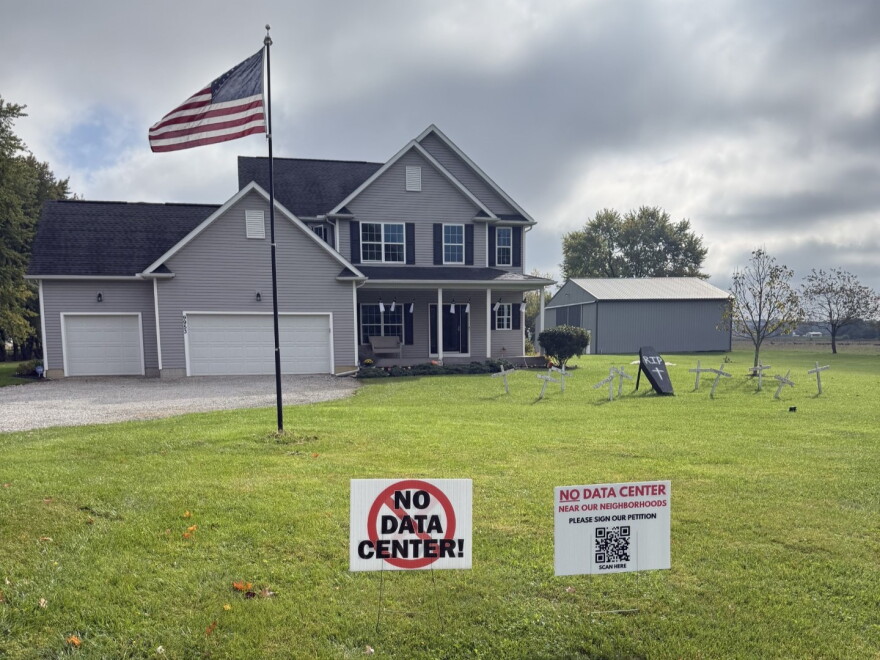 Two signs in front of a large residential home on a rural plot read "No Data Center!" and "No data center near our neighborhoods, please sign our petition," with a QR code.