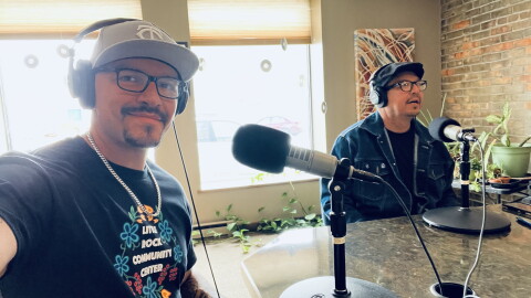 Two men in a radio studio sitting behind microphones. One is holding his arm out to take the selfie.