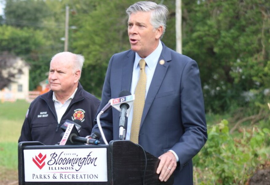 Darin LaHood speaks at the Bloomington Parks & Recreation podium as Mayor Dan Brady stands behind him in his fire department jacket.