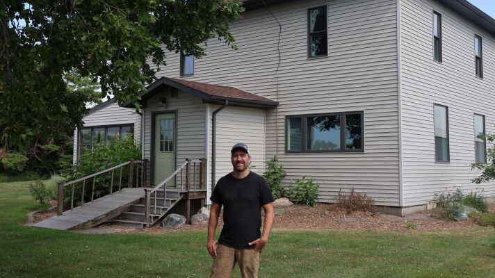 A man in his forties wearing a black shirt stands in front of his home, which has stood for over a hundred years.