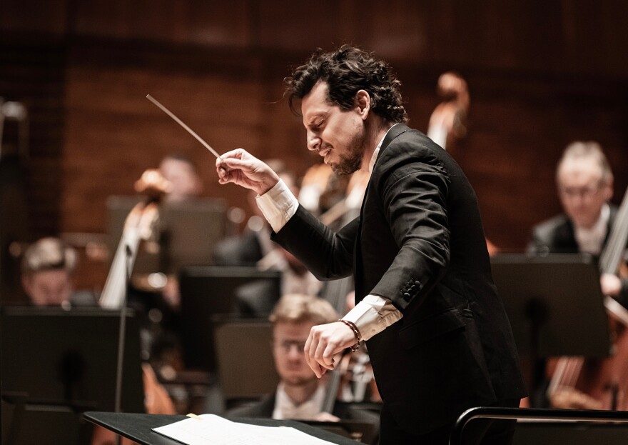 a person with eyes closed in a dark suit holding a conducting baton while musicians play in the background