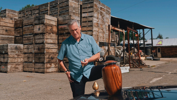 Bruce Campbell in a still from Ernie & Emma, smoking a cigarette while looking down at an urn placed on the hood of a car. There's a golden pear as the hood ornament.