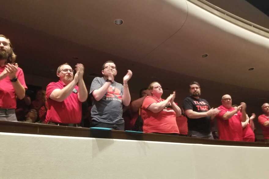 A row of people, mostly wearing red shirts, stand clapping at the edge of an auditorium balcony seating. More people can be barely seen in the darkness behind them, also clapping.