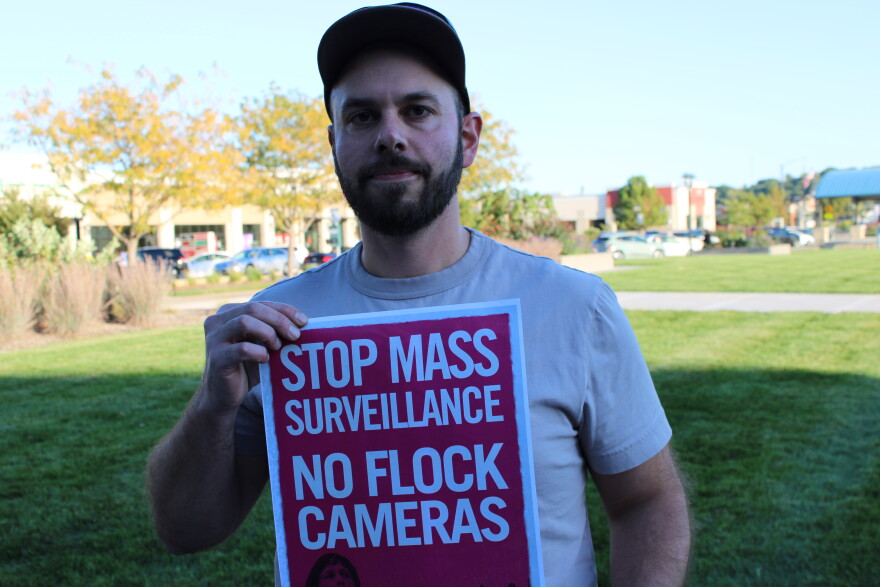 A man wearing a baseball cap holds a sign that reads “STOP MASS SURVEILLANCE NO FLOCK CAMERAS.”