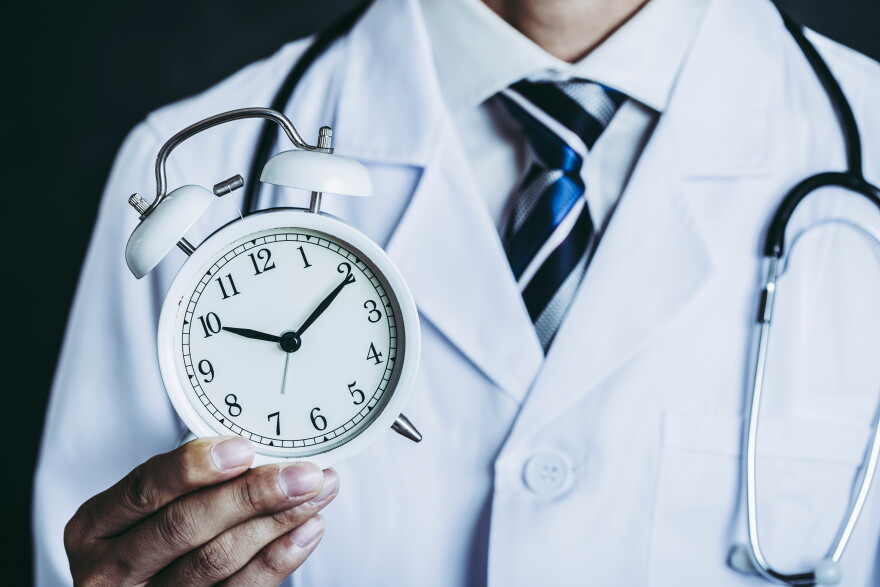 A doctor in a white lab coat and blue-striped tie, a stethoscope around their neck, holds up a white alarm clock showing about 10:10.