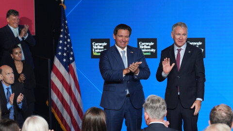 Two men wearing suits wave as they stand on a stage next to an American flag, flanked by people clapping. 