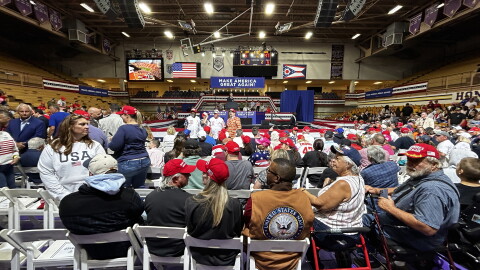 A large crowd of people seated in a high school gymnasium with a large sign in the back that reads "Make America Great Again."