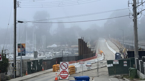 Fencing blocks off the right lane of a bridge while the left side is open. Sailboats float in water on the left side of the image.