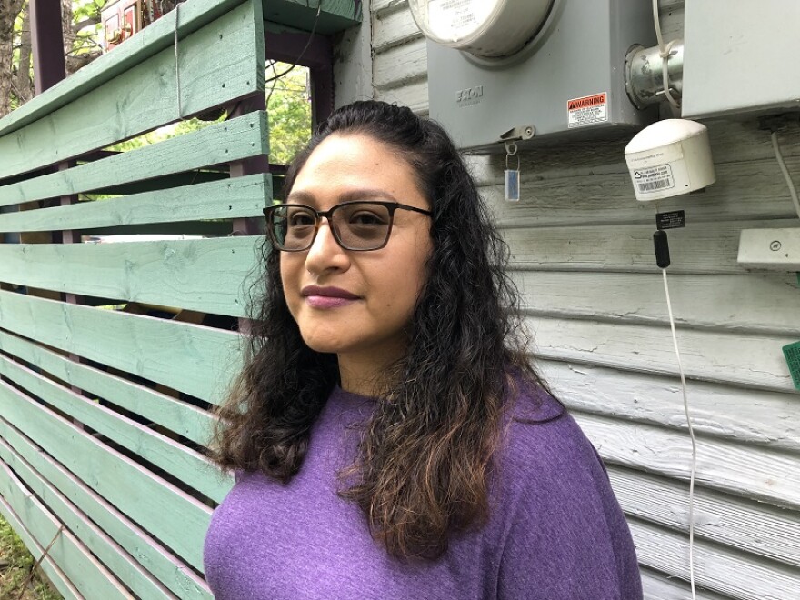 A woman in a purple shirt stands outside of a house in front of the electric meter. Next to the meter is a small, white cylinder that is an air quality sensor.