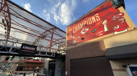 An outdoor photo shows a large red and yellow mural on top of a brick building. It shows depictions of football players dressed in Kansas City Chiefs jerseys, a Super Bowl Trophy and other Kansas City Chiefs graphic elements. Next to the building is an outdoor venue with a sign that reads "Kansas City Live."