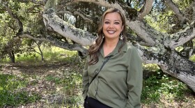 A woman with long hair, dark pants, and an olive green blouse smiles while standing in front of a large tree.