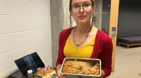 Young woman with her hair pulled back is holding a silver pan of baked cake. She is wearing round rimmed glasses, a bright red cardigan over a yellow shirt. 