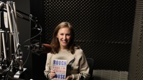 A smiling woman sits in a soundproof radio studio beside a microphone, holding up a book titled “Rough House.”