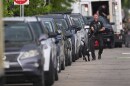 An officer walks a black police K9 along cars parked on the side of a road. In the background, you can see more law enforcement.