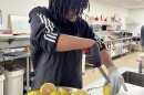 A teenager cuts grapefruits in an industrial kitchen. He's wearing and apron and gloves.