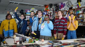 Seven performers stand behind an office desk with smiles on their faces while sum of them hold shucked ears of corn.