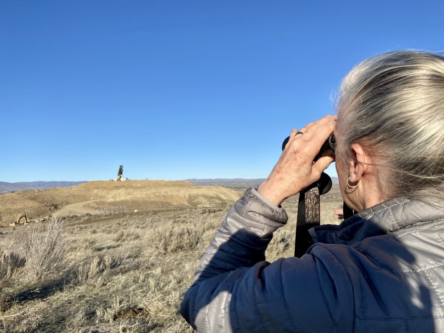 A woman in a gray jacket holds binoculars. She's looking at a landfill.