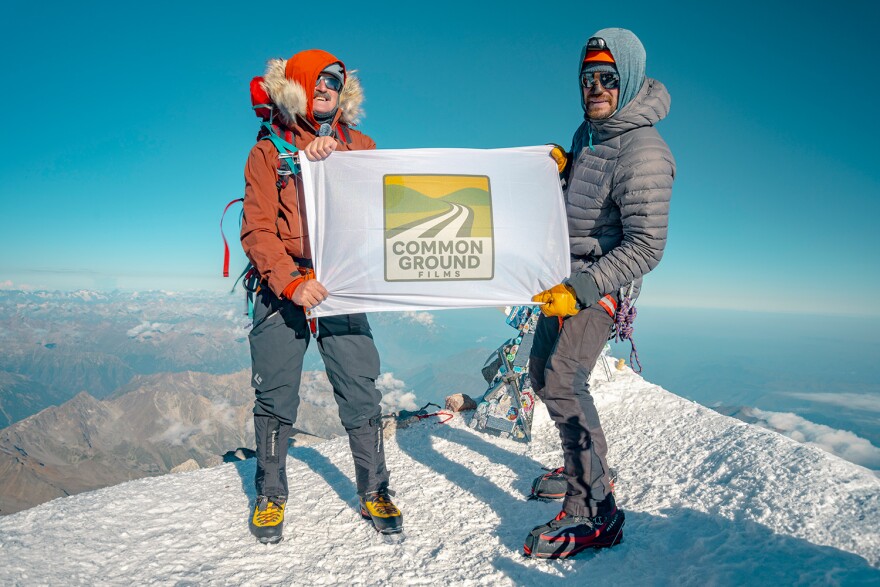 Two men on a snowy mountain peak in climbing gear hold a banner between them that says "Common Ground Films"