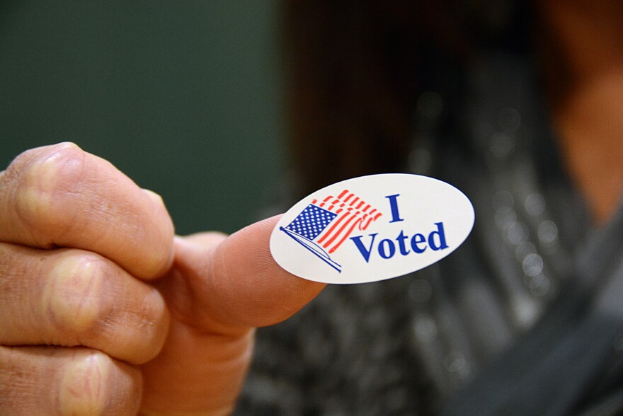 A hand holding up an I Voted sticker with an American flag.