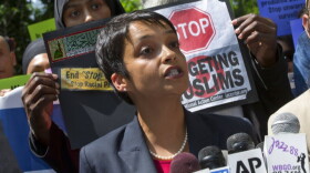 A young woman with short dark hair and dark complexion in a gray blazer and pearl like necklace speaking behind microphones with people behind her holding up signs about targeting muslims. 