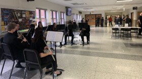 A small group of high schoolers dressed in black smile and play instruments. In the background, parents file into an auditorium