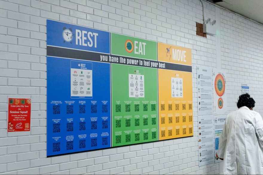A large, three-section informational board titled "REST, EAT, MOVE" hangs on a white brick wall, with the tagline "you have the power to feel your best." The "REST" section is blue, the "EAT" section is green, and the "MOVE" section is orange, each featuring columns of individual QR codes for scanning. To the right, a person wearing a white lab coat stands with their back to the camera, looking at adjacent posters.