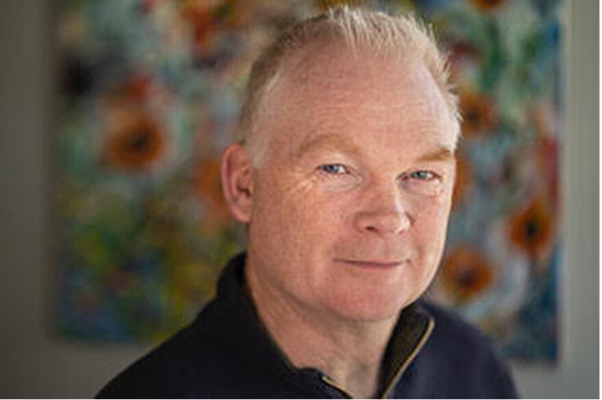 Graeme Reid poses for a headshot photo. He’s indoors. Behind him is an out-of-focus piece of art on the wall.