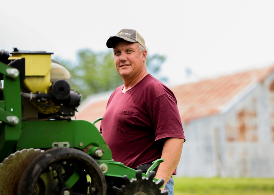 A man wearing a baseball hat, a maroon t-shirt, and jeans stands behind a green piece of farm equipment. 