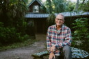 A man in a plaid long sleeved shirt is sitting outdoors on top of a decorative tiled table that's in front of a forrested cabin