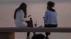 Two women sit on a bench at sunset, enjoying some snacks and looking at their phones