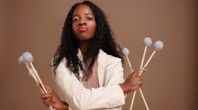 African American woman with long black wavy hair and white jacket holding six percussion mallets in crosses arms