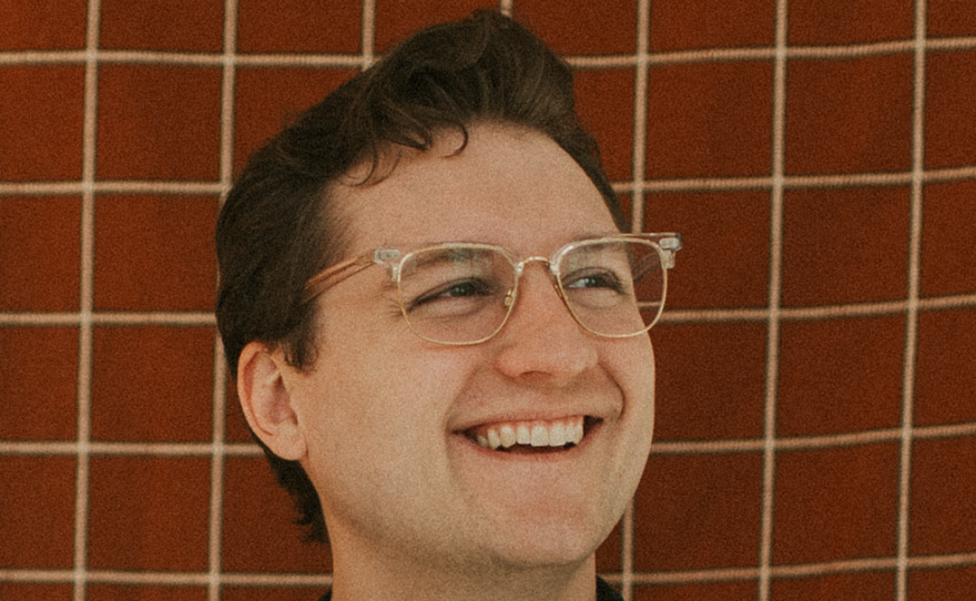 A smiling man with short brown hair stands in front of a tiled wall. He is wearing glasses and a black sweater and looking up.