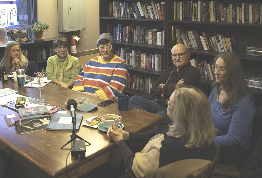 A group of people sit around a large, rectangular wood table with drinks and books atop it. One older woman holds the group's attention