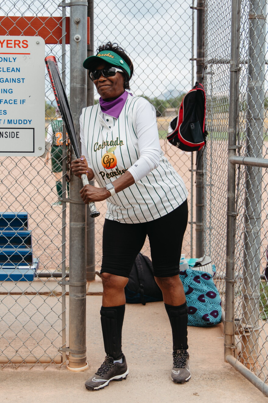 A woman leans up against a metal fence wearing a green visor and a white baseball jersey that reads "Colorado Peaches." She's holding a silver baseball bat in her hand and smiling. Behind her is a dugout and a baseball field.