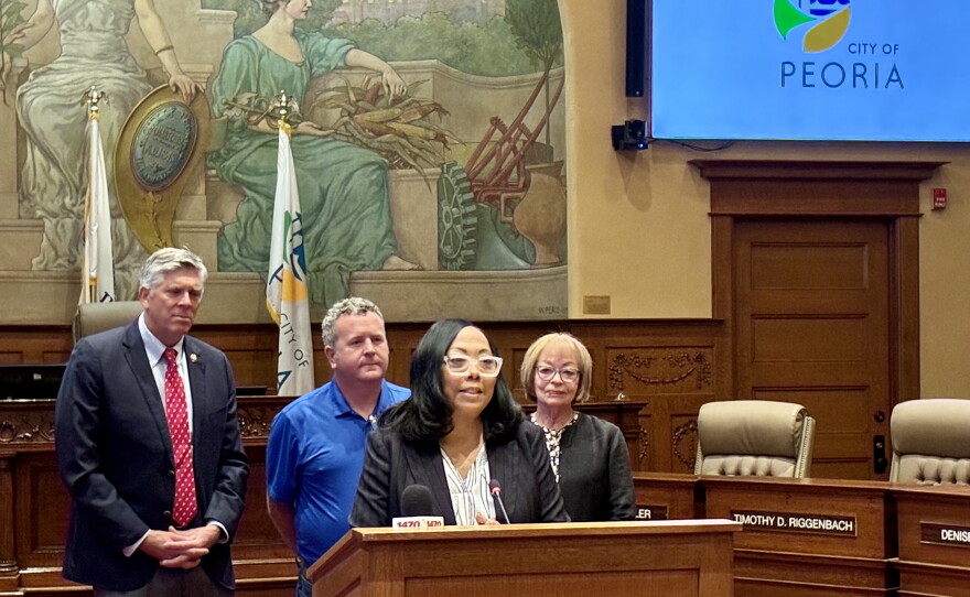 Four people stand at a podium in a formal room with a mural and city flags behind them. A woman speaks at the microphone, while three others stand behind her. A screen shows the City of Peoria logo.