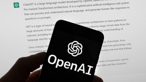 The OpenAI logo is seen on a mobile phone in front of a computer screen which displays output from ChatGPT.