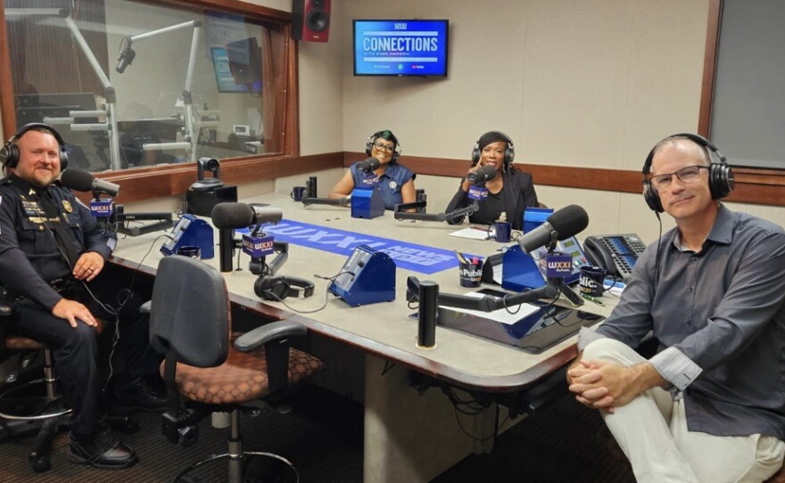Four smiling people sit around a table in a radio talk studio: a man front left has short light brown hair and a light brown mustache and beard and is wearing a police uniform; a woman back left has short blue hair and is wearing glasses and a blue top; a woman back center has long black hair and is wearing a black top and blazer; a man at right has very short dark hair and is wearing glasses, a grey button-down shirt, and white linen pants