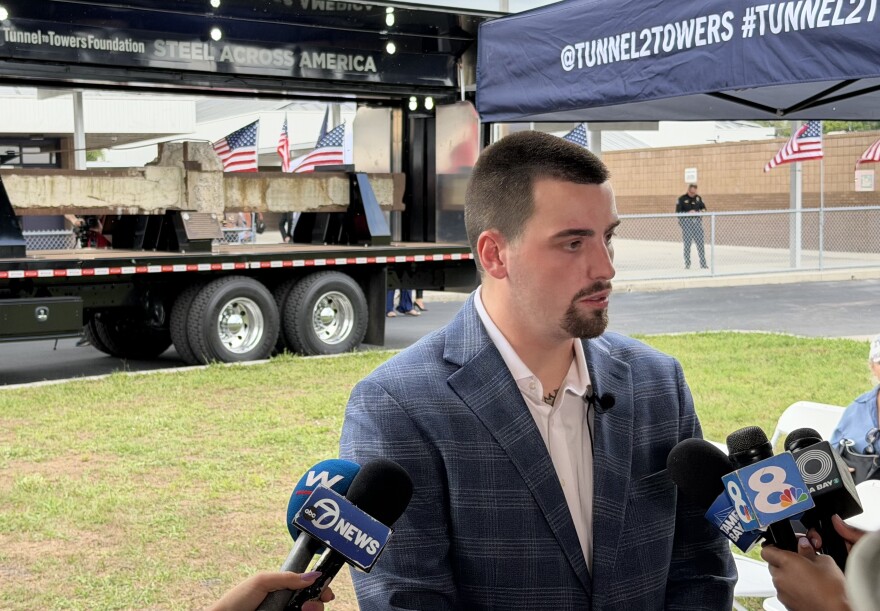 A young man in a white shirt and blue blazer is interviewed by reporters by a truck carrying the WTC beam visible in the background. 