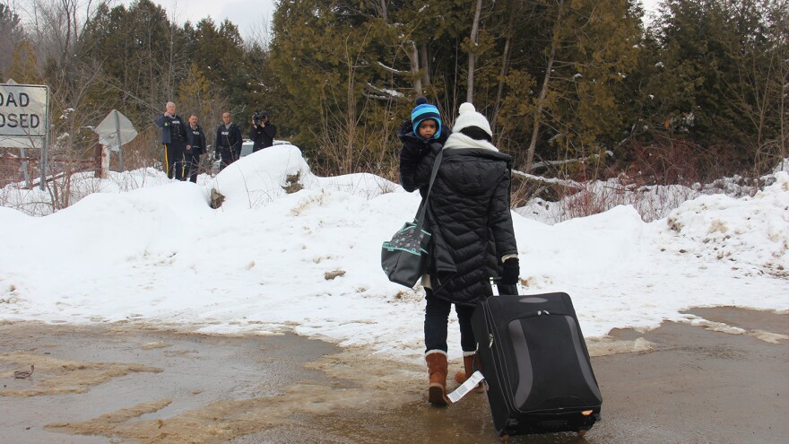 A woman traveling alone with her infant, seeming to understand that she will be arrested, walks toward Canadian police on the far side of the border.