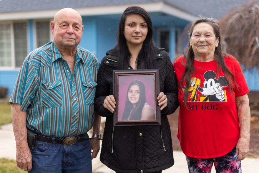 A young woman stands between older people, holding an image of a young teen girl.