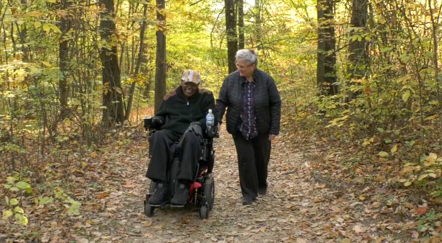 Man in wheelchair and woman walking use forest trail.