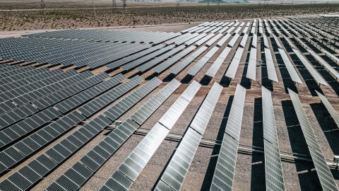 This is an image of a large-scale solar farm with rows of solar panels angled toward the sun on a sunny day. Powerlines and mountains are in the background. 