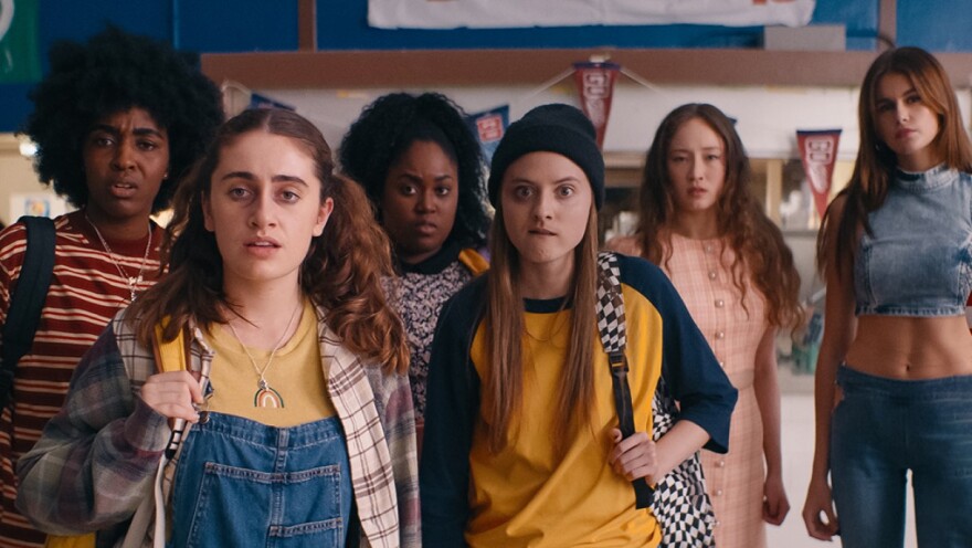 Six female high-school students look quizzically at something out of frame as they stand in the hallway near the school's trophy case.