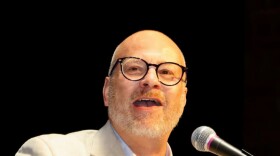A man in a cream-colored suit jacket and glasses stands behind a wooden podium, looking to his right at the camera